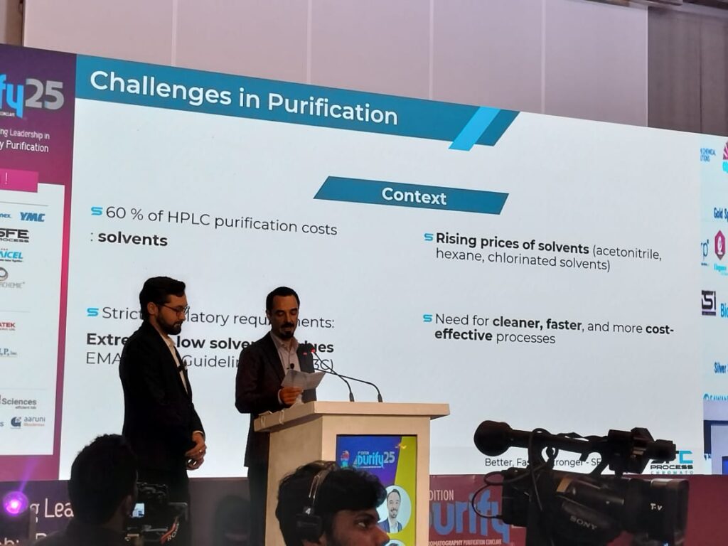 Speaker at the podium in front of the Challenges in Purification slide on solvent costs and regulations