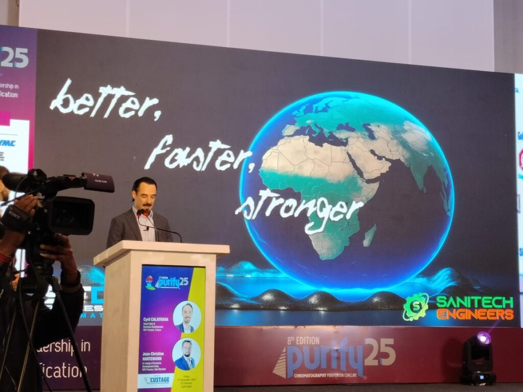 Speaker at the podium beside a glowing globe screen reading 'better, faster, stronger' at Purify 25