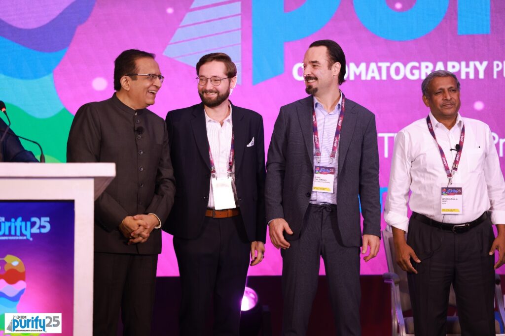 Four delegates standing beside the podium in front of the Purify 25 chromatography stage backdrop
